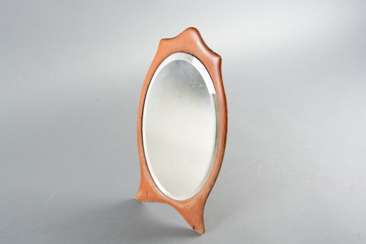 Ovalt speil med treramme. Rammen følger speilets form med tre avrundede spisser på toppen og to avrundede føtter nederst. På baksiden er det festet et stativ av metall.
