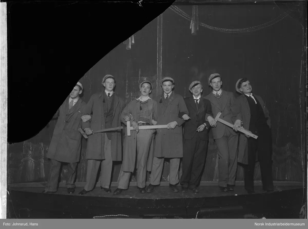 Kveldsunderholdning i teateret av Juventas. Syv gutter oppstilt på scenen med teater rekvisitter i hendene og russeluer på hodet.