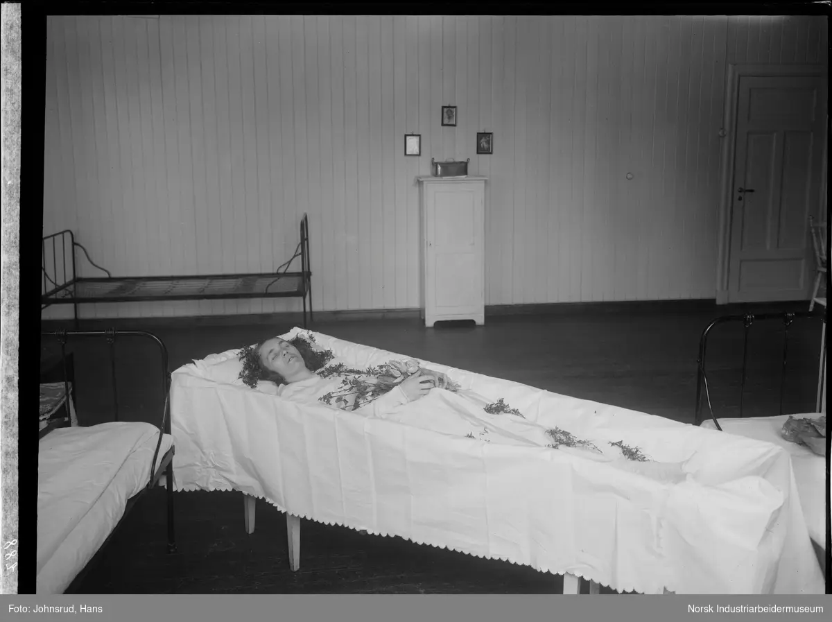 Post mortem fotografi av kvinne liggende i åpen kiste. Kvinnen er kledd i hvite klær og har blomster dekorert på og rundt seg. Kisten står i et rom med flere senger, muligens ett sykehusrom.