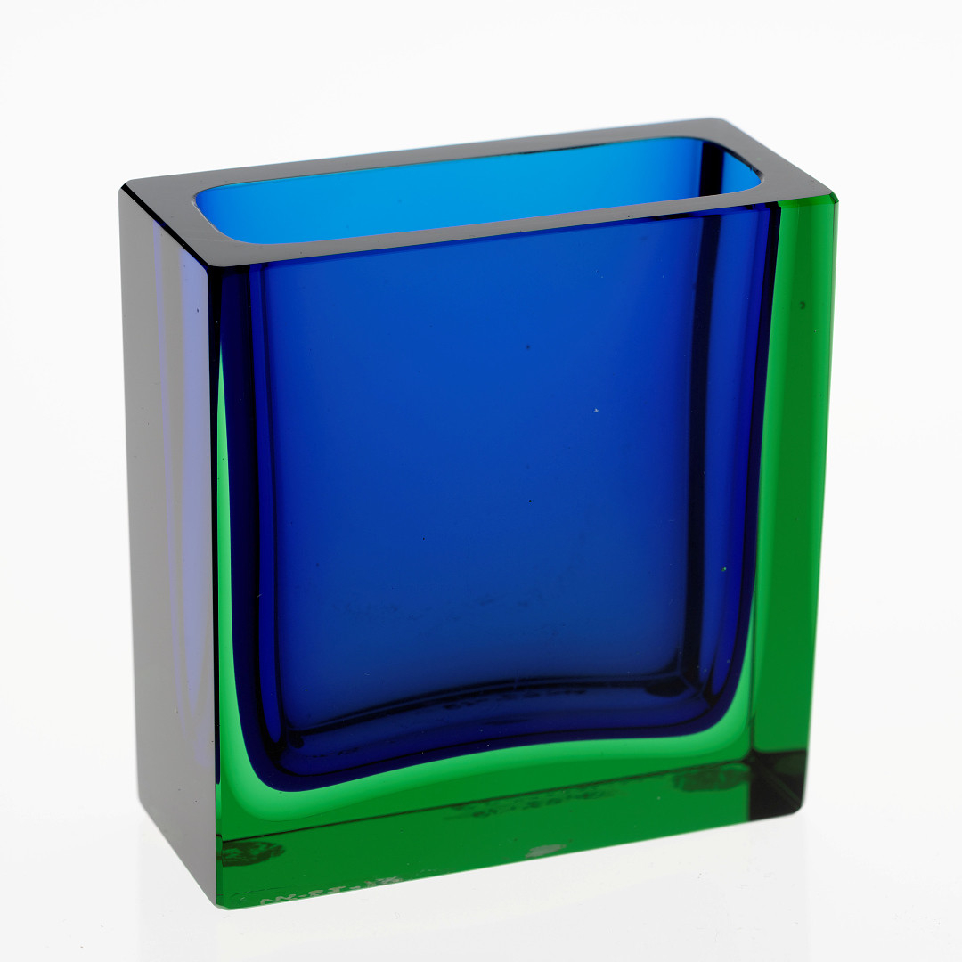 Rektangulært sigarettbeger i gjennomskinnelig overfangsglass med planslipte sider. Blåfarget indre kjerne, mens de ytre sjiktene er i grønnfarget glass. 
