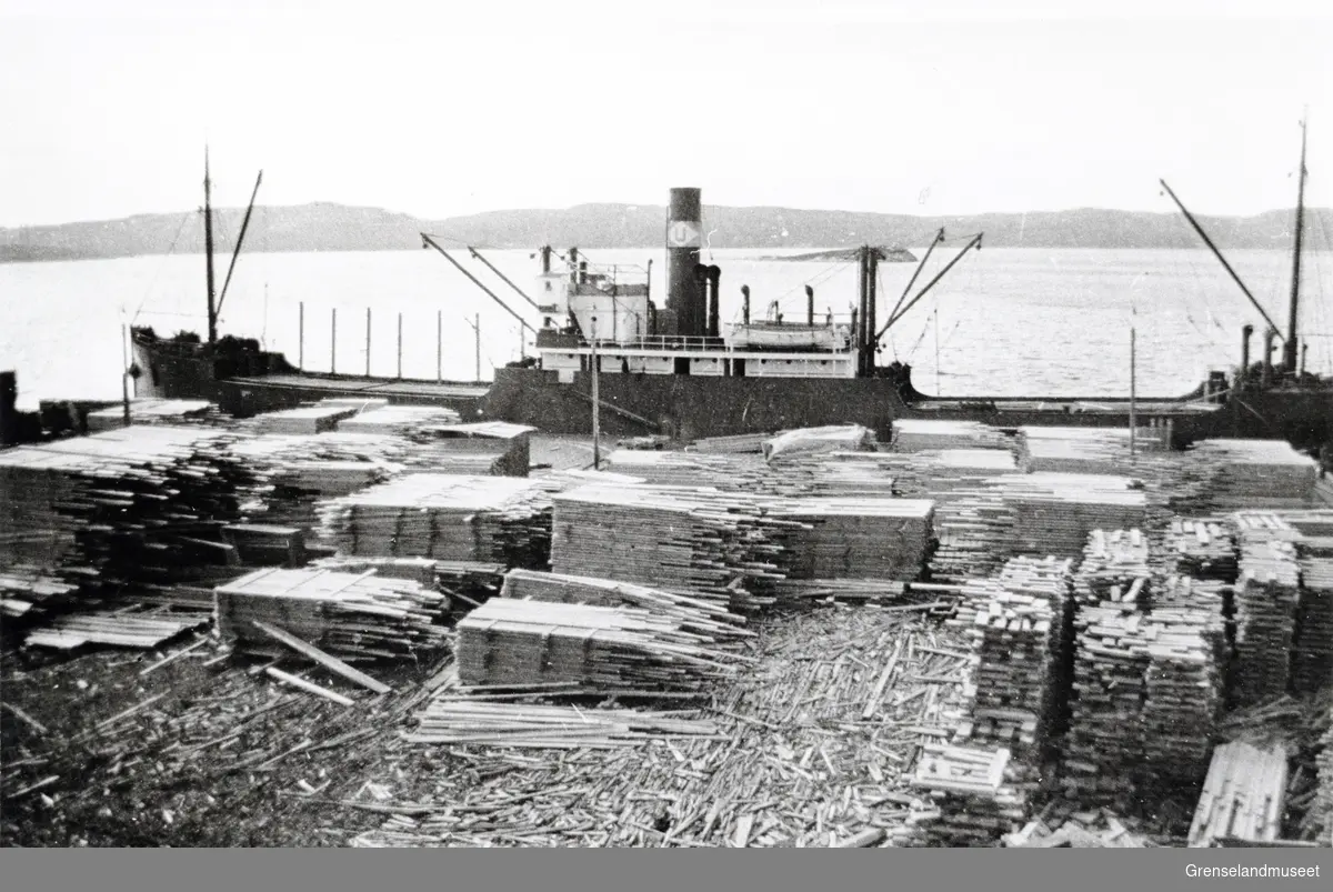 En båt ligger til havn ved sagbruket til Pasvik Timber på Jakobsnes. Kaia er full av plankestabler. 
