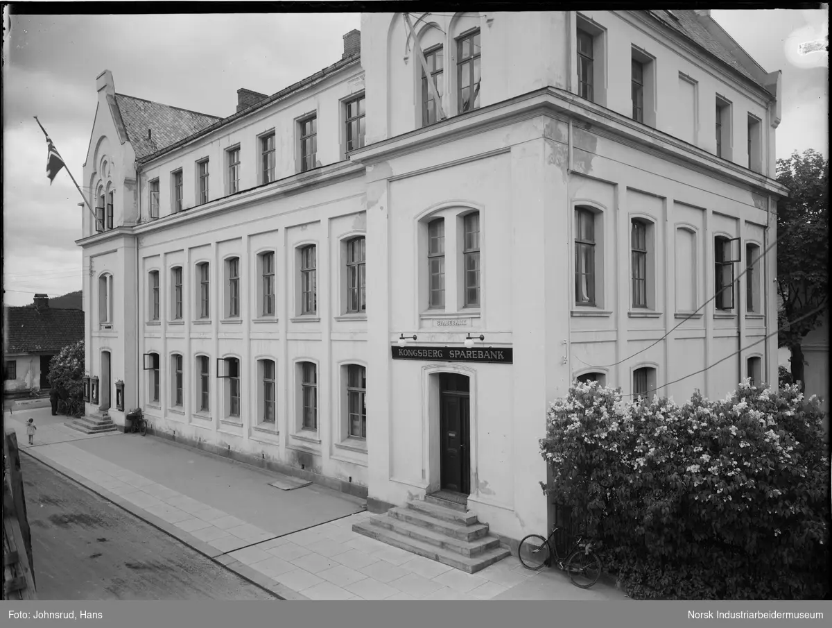 Bygning i mur for Kongsberg Sparebank. Jente gående i gaten på utsiden av banken. Sykkel lent inntil gjerde på siden av bankbygning.