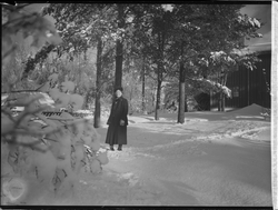 Kvinne kledd i vinterkåpe, votter og hatt stående i hage på 