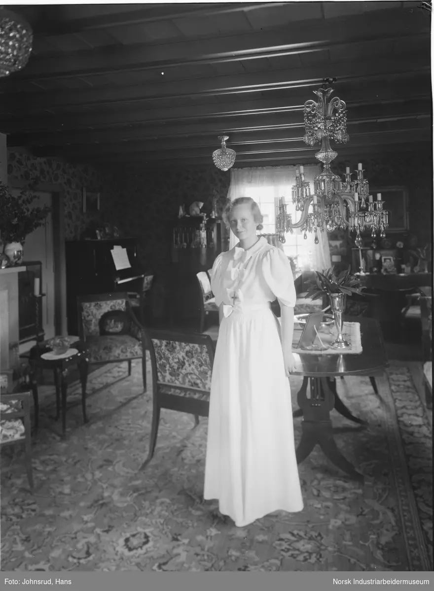 Kvinne i lys kjole oppstilt i stue. Stuen er møblert med to spisestuemøblement og peis. Lysekrone hengende fra taket.
