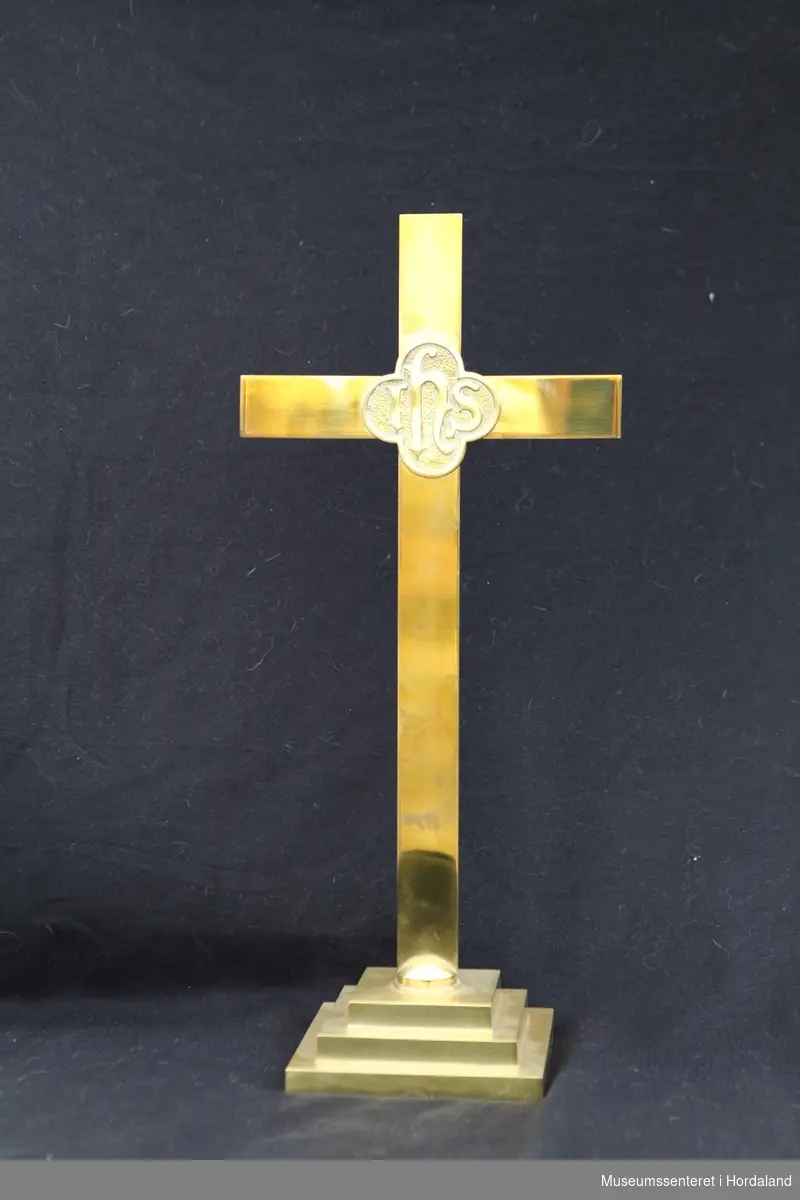 Alterkors i messing. Inngavert på midten av korset står "IHS" i en firearmet rosett. Korset står på en kvadratisk fot.