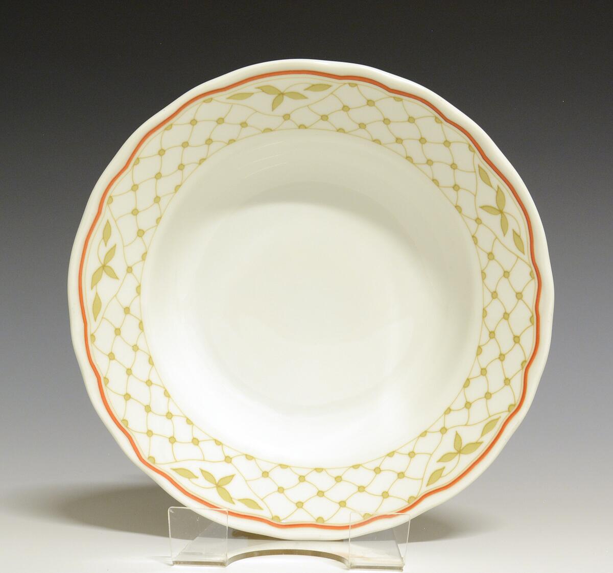 Dyp tallerken av porselen med hvit glasur. Bølget kant. Fanen dekorert med oransje kantstrek og et nett i gult med fem utsparte felter med blader i.