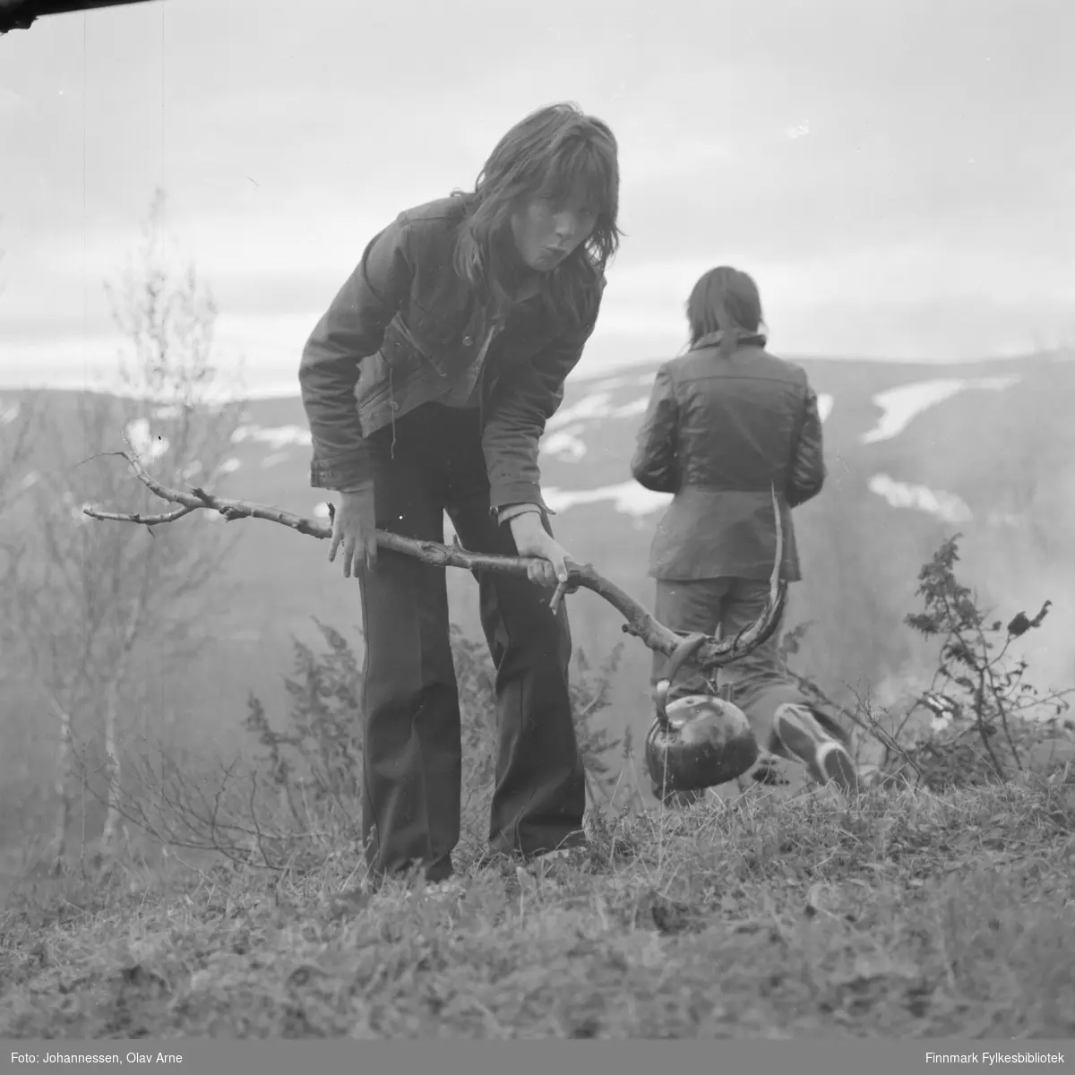 Ungdom på tur i Finnmark

Et av ungdommene ser mot kamera med en grimase og holder på en stor kvist med en kaffekjele hengende på. I bakgrunnen ser man en anne person i gummistøvler
