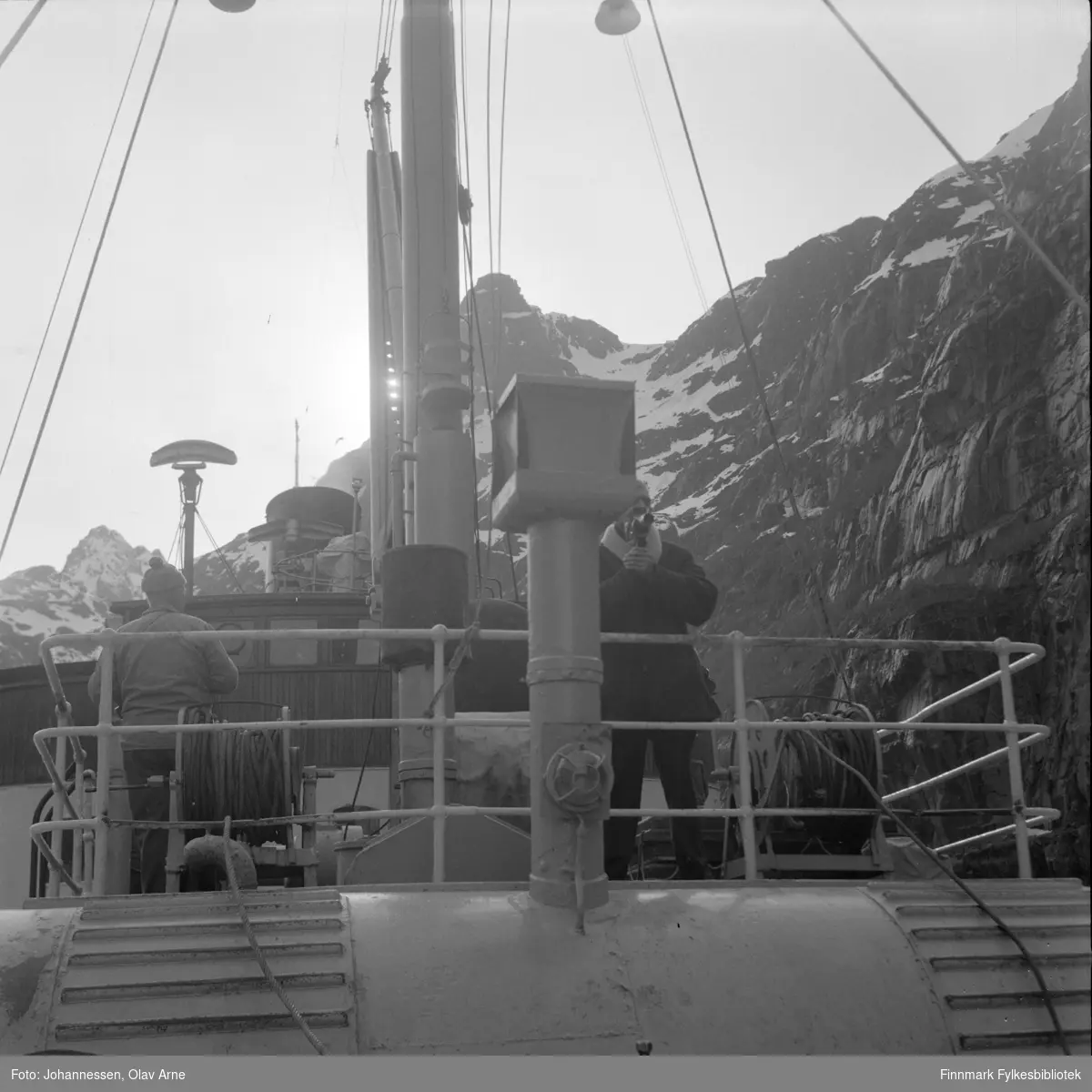 Ombord i DS "Lofoten" som var i hurtigrutefart for Vesteraalske fra 1931 til 1964. Bildet er tatt i Trollfjorden i Nordland.

Mannen til høyre holder på et film kamera og man kan se fjell i bakgrunnen.