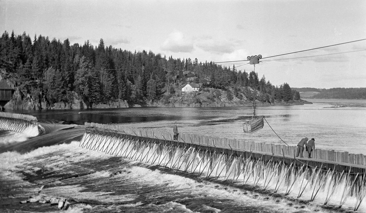 Bygging av såkalt fermettedam ved Mørkfoss i nedre del av Glomma høsten 1914.  Dette er en forholdsvis lett, men robust nåledamtype, der nålene holdes på plass av stålbukker som er hengslet til botnelementer på en måte som gjør det overkommelig å demontere dem om det skulle være påkrevet i reparasjonsøyemed.  Fotografiet viser konstruksjonen sett i et skrått perspektiv fra motstrøms side.  Øverst på «baksida» av de nevnte bukkene var det montert gangbruer, som fløterne kunne arbeide fra når dammen skulle settes eller åpnes.  Da dette fotografiet ble tatt sto det tre karer på denne gangbrua, med blikkene vendt mot vannet på motstrøms side.  Midt i elveløpet var det en åpning, hvor vannet fosset ut.  Over konstruksjonen ser vi ei taubane med løperkatt, som åpenbart ble brukt til å heise på plass damelementer.  På motsatt side av elva ser vi et berglendt terreng med en del skog, blant annet et lite våningshus (sentralt i bildeflata) og noe som kan se ut til å ha vært et redskapshus (ved venstre bildekant). Dammen på fotografiet ble sannsynligvis demontert vinteren 1924, for fra påfølgende vår var det den da nye dammen ved Solbergfossen som regulerte Øyeren. 

Den første fermettedammen ble konstruert av den franske ingeniøren Poirée i forbindelse med kanaliseringa av Mainz så tidlig som i 1831.  Dette anlegget ved Mørkfoss kan ha vært det første i sitt slag her til lands.  Denne dammen må ha virket som en terskel i vassdraget, i og med at den hevet vannspeilet oppover mot Øyeren, og dermed gjorde det enklere å få tømmeret fra Øyeren ned til Mørkfossen uten at det strandet på steinskjær og grusører, samtidig som det ble mulig å gå ned i dette området med små slepebåter. Mørkfossen fikk sin fermettedam ved inngangen til 1860-åra som ledd i et kanaliseringsprosjekt der kanaldirektør Balle Røyem (1813-1874) hadde vært sentral i utredningsarbeidet - de fanen «Oplysninger».