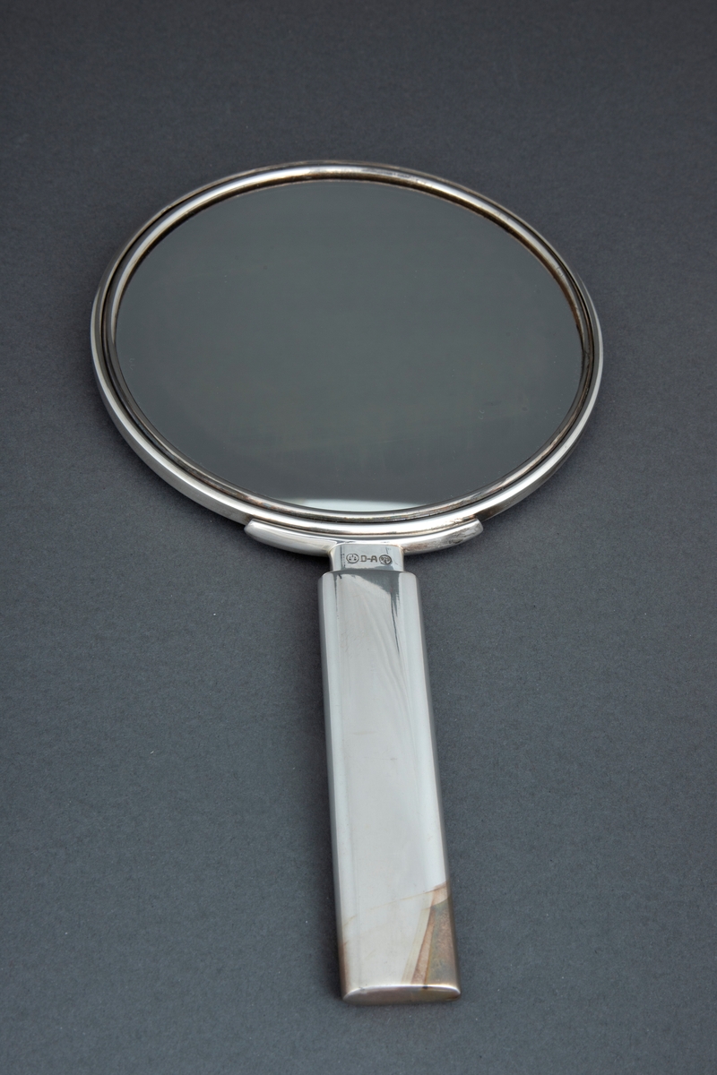 Sirkelrund speil med håndtak. Speilet er innfattet i sølv og har en lett konveks bakside med sirkel i relieff. Flat, rektangulært håndtak i sølv med avrundede kanter.
