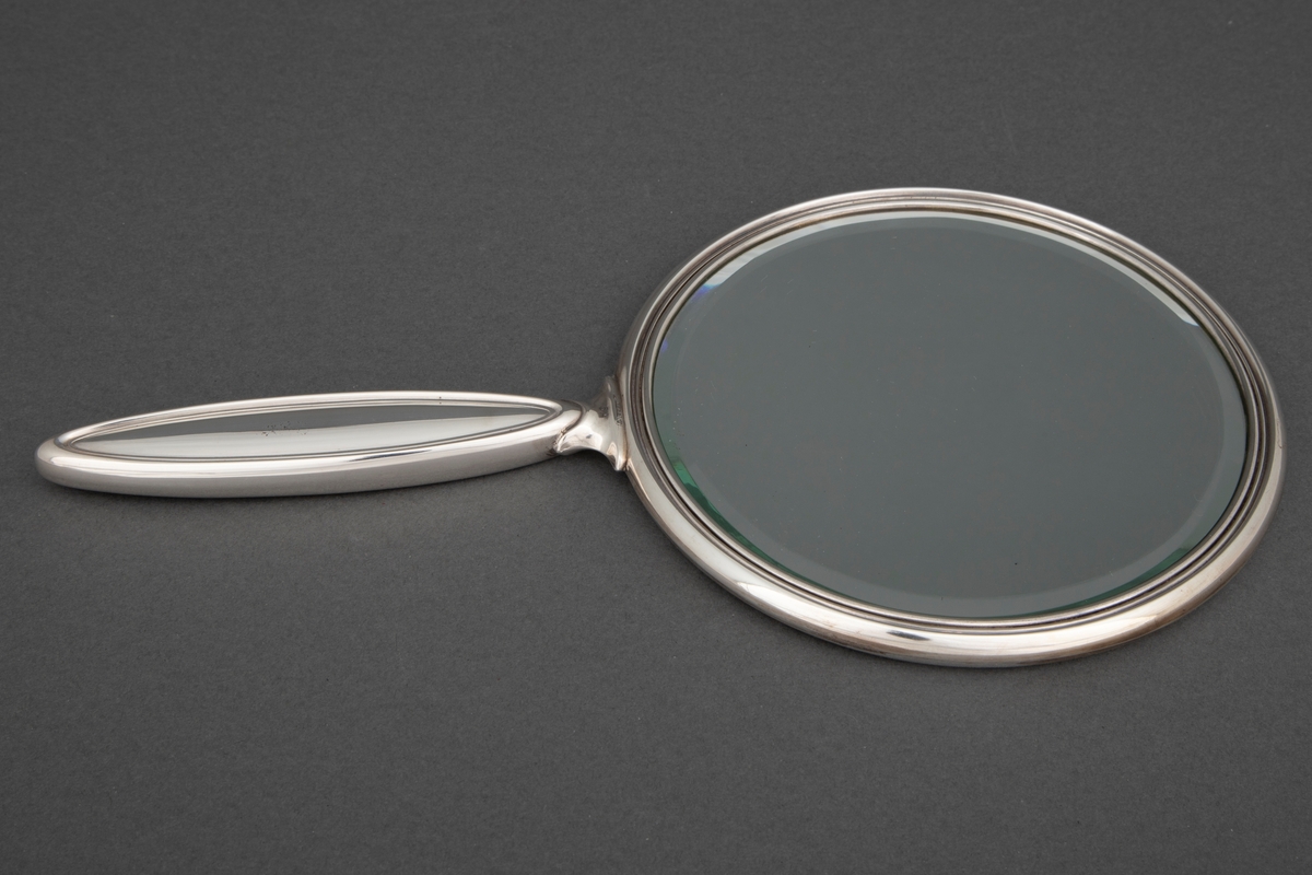 Sirkelrund speil med håndtak. Speilet er innfattet i sølv og har flat bakside med en profilert rand. Håndtaket har form som en avlang spiss oval med profilerte render på for- og bakside.