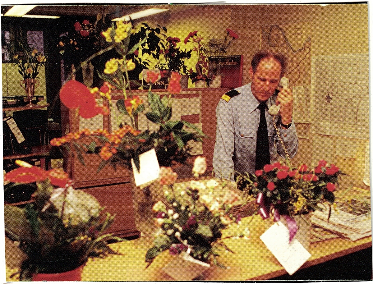 En uniformert politimann snakker i telefonen stående bak et skrivebord. Mannen er omgitt av mange blomsterbuketter.