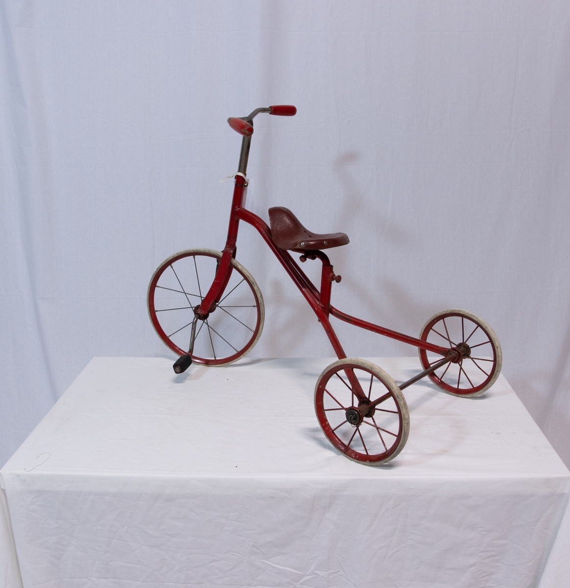 Trehjulsykkel m/rødmalt sykkelramme i stål. To mindre bakhjul, et stort forhjul. Eiker i lettmetall. Hard gråhvit gummi på hjulene. På pedalene tredd to gummi fotstykker, riflemønster. På styret
to sylindriske trehåndtak, rødmalt.