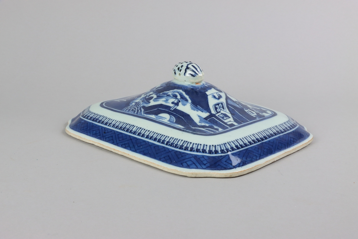 Square form with chamfered corners and slightly domed shape, on top a knob in form of a fruit. On the lid landscape scenes of pagodas, buildings,  bridges, figures with parasolls,  gardens and waters. The edge of the lid is decorated with a wide dark blue border with a criss cross pattern. All decorations in blue underglaze. The inside of the lid without decoration.