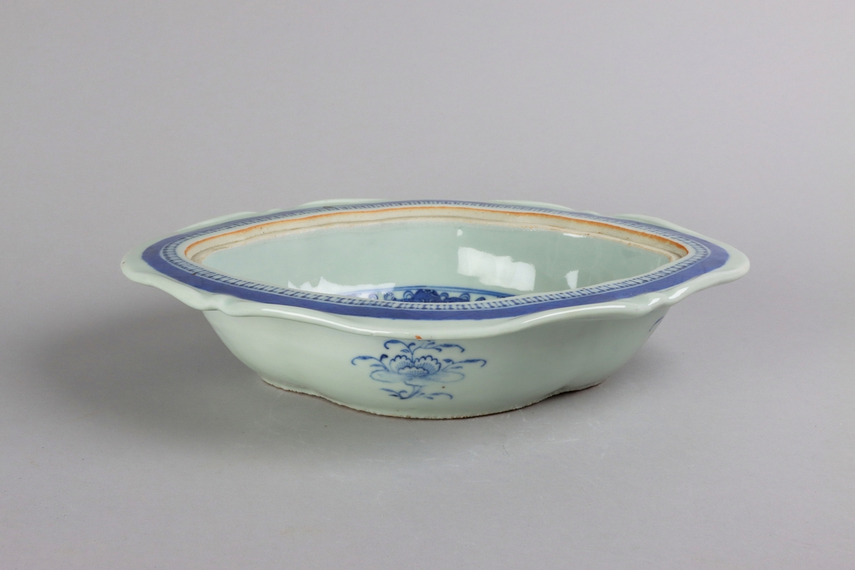 Oval broken form with a wide flat rim, decorated with a dark blue border with criss cross pattern. The inside of the dish slightly everted and undecorated. The well surrounded with a border of a criss cross pattern and rectangular reserves filled with symbols of good fortune. Centered in the well landscapes scenes with pagodas, buildings, waters, bridges and gardens. The outside of the dish is decorated with rose branches. All decor in blue underglaze. The base of the dish without glazing.
