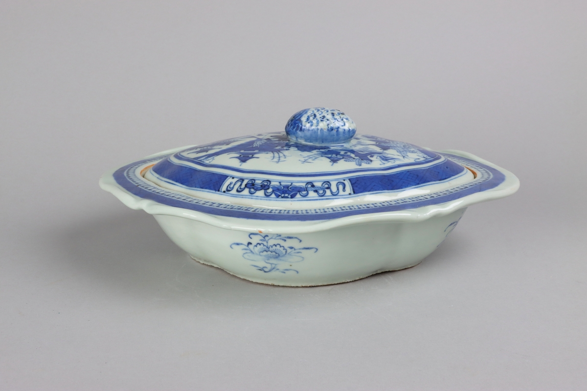 The lid with an oval slightly domed form, on top a knob shaped as a fruit. On the lid landscape scenes of pagodas, buildings,  bridges, figures with parasolls,  gardens and waters. The edge of the lid is decorated with a wide dark blue border with a criss cross pattern and reserves filled with symbols of good fortune.. The outside of the dish is decorated with rose branches. All decor in blue underglaze. The base of the dish without glazing