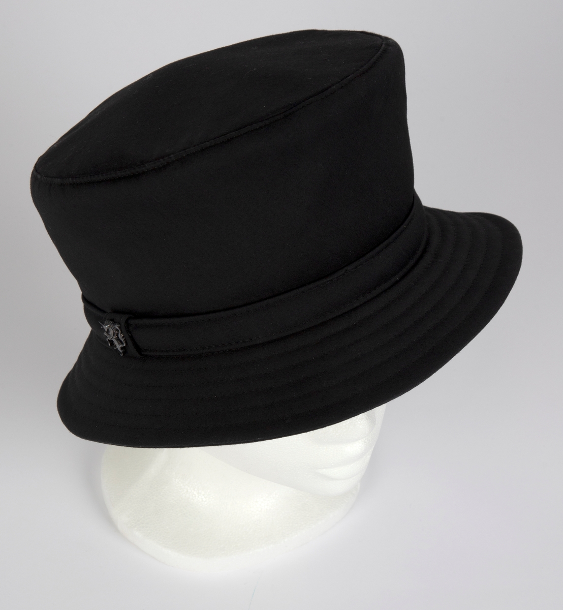 Relativt høy hatt av kraftig, kypertvevd, mercerisert bomull. Flosshatt-type, med tilnærmet sylindrisk pull, lett utsvinget øverst, noe høyere foran og med flatt, sirkulært toppstykke. Smal brem, som skråner nedover, stukket med tettstilte, parallelle sømmer. Pull og brem har søm midt bak. Smal, kantstukket hattebånd, med skjult skjøt under en "løkke" av hattebåndet, midt på høyre side. Denne "løkken" er dekorert med designerens logo, en steilende enhjørning i antrasittgrått, blankt metallrelieff. Innvendig fôret med marineblått, blankt silkestoff, foldelagt inn mot et sirkelformet midtparti, avgrenset mot foldene med en smal, gyllen silkerulour. Bredt svettebånd i kraftig bomullsrips. Hatten har også mellomfôr i pullen, antagelig med et tynt lag Polyester.