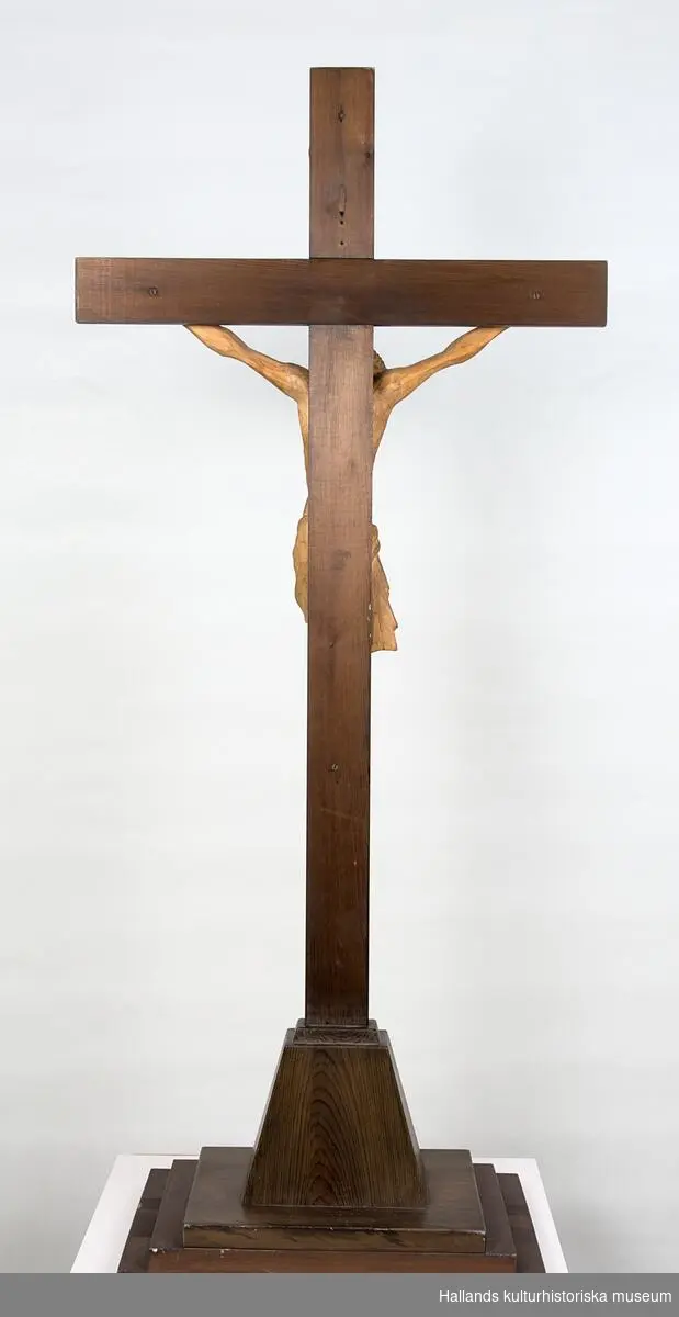 Skulptur av trä. Plattform och kors av mörkbetsat trä (furu). Jesusfigur handsnidad av ädelträ (troligtvis valnöt). Ovanför Jesusfiguren en skylt: "INRI". Svårläst signatur i blyerts på figurens rygg: "NJ??? 15/11 29".