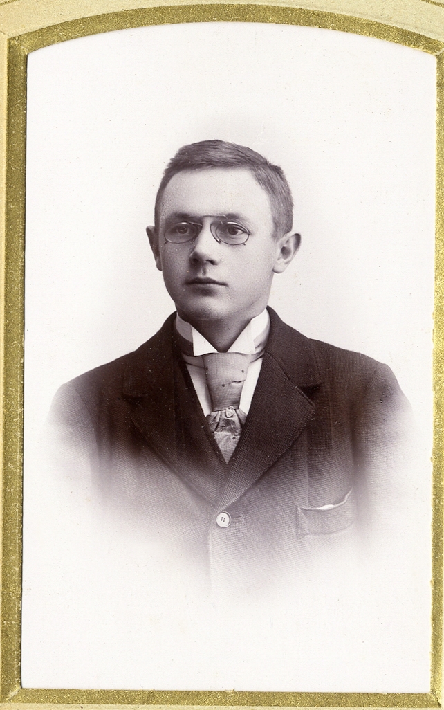En ung man med pincené, klädd i mörk kostym, stärkkrage och ljus slips med sköldformad slipsnål/pin.
Bröstbild, en face. Ateljéfoto.