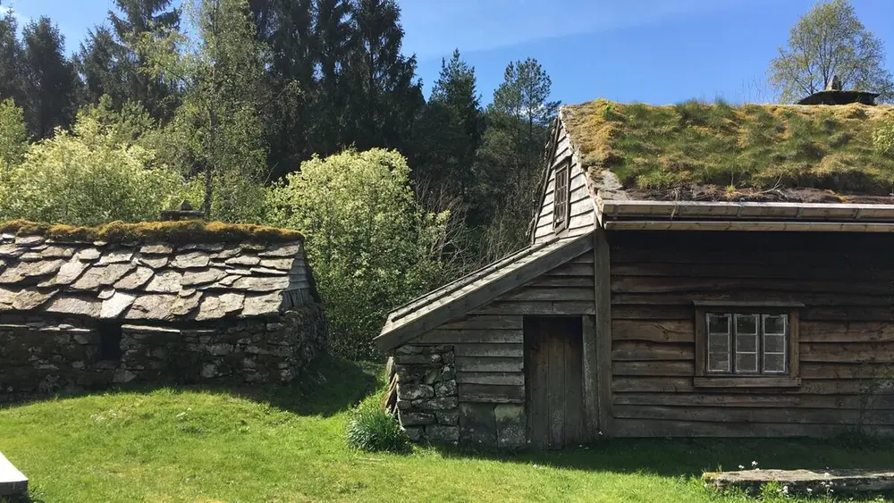 Hus i museumstun med torvtak, skykkjer og villskifertak i sommersol