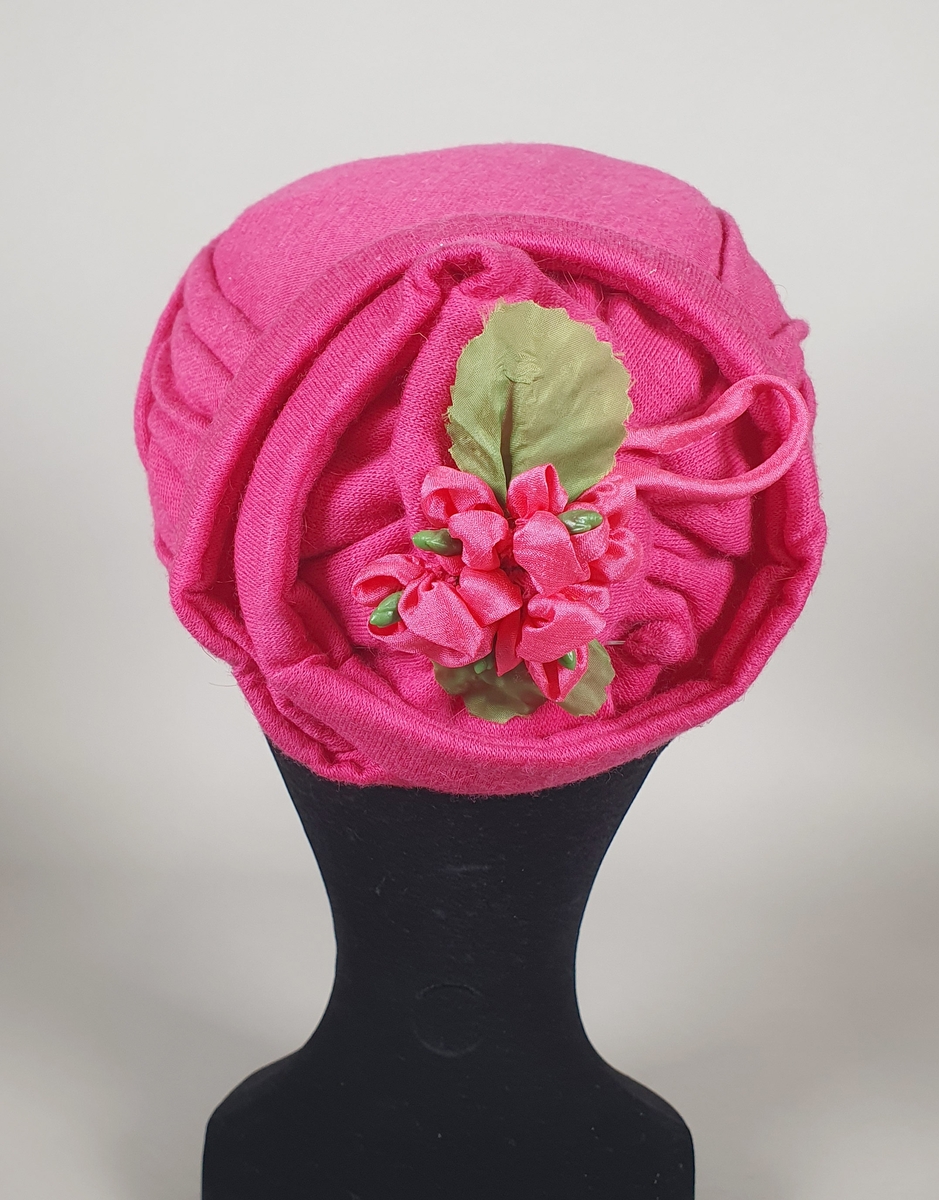 Rosa sommerhatt av bomull, uten brem. Stoffet er brettet rundt pullen, og bak er det et rundt felt med rosett og hattenål. Hatten er fôret med hvit kunstsilke og har et hvitt ripsbånd rundt kanten.
