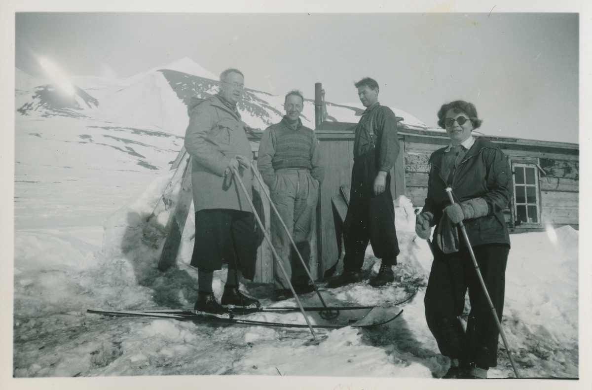 Tre menn og ei kvinne foran hytte. Bildet er del av fotoalbum SVB 2668