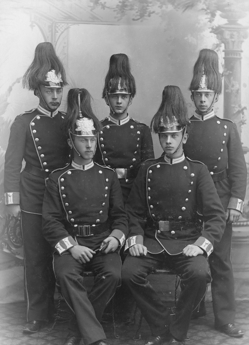 Ur grosshandlare Oskar Nordbloms fotoalbum. Fem unga män i Trängkårens uniform m/1885 med plymförsedda kaskar (pickelhuvor). Trängbataljonen var det första trängförbandet och uppsattes 1885 i Stockholm med ett kompani för allmän trängtjänst och ett för sjukvårdstjänst. 1891 delades den upp i Svea trängbataljon och Göta trängbataljon; den senare förlades till Karlsborg. Trängtruppernas uppgift är att utbilda personal till underhållsförband, tillhandahålla förnödenheter, reparera skadad materiel, föra undan och vårda sårad och sjuk personal samt vid krig mobilisera dessa.