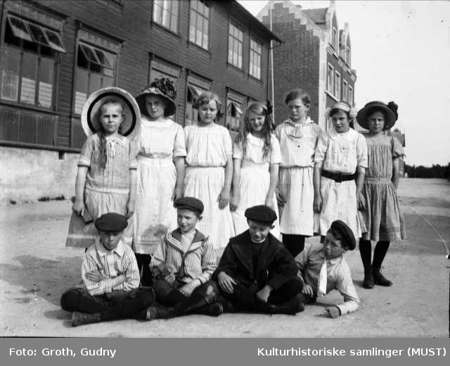Gruppeportrett utendørs. Syv jenter og fire gutter i finklær foran Platou skole i Birkelandsgaten (tidligere Frk. Rings skole).