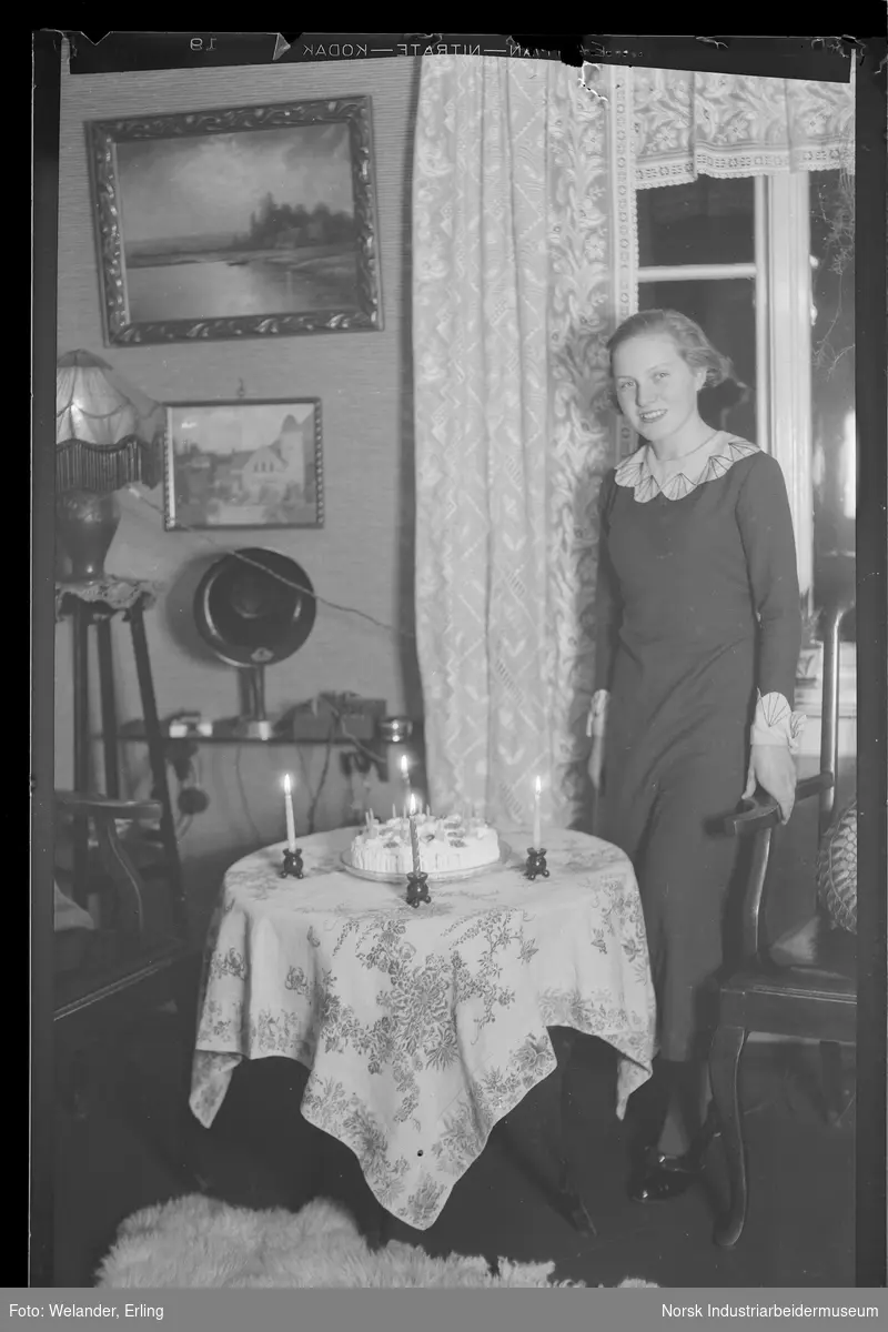 Kvinne stående foran bord i stue. På bordet står en bløtkake, lys stående på bordet rundt kaka. Maleri og fotografi av Rjukan kirke i bakgrunnen.