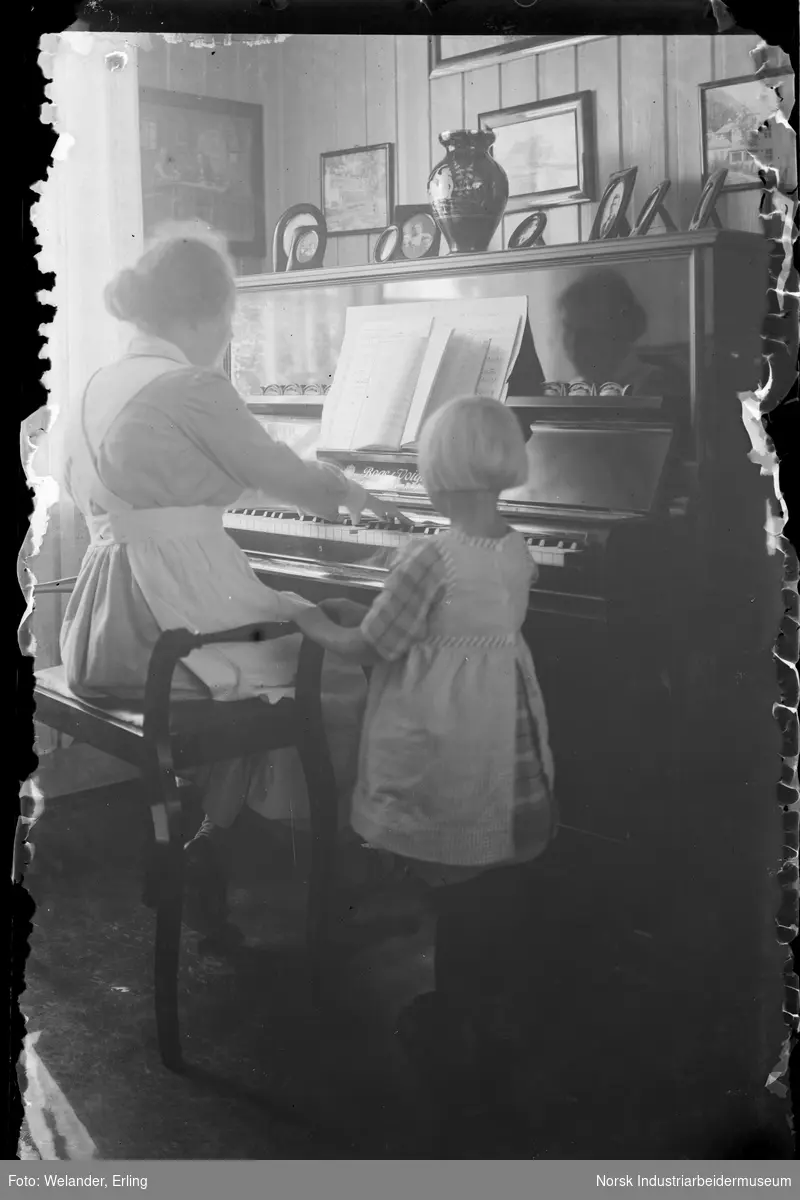 Kvinne spiller på piano i stue. Barn står ved siden av å ser på. Fotografier på veggene og på toppen av pianoet.