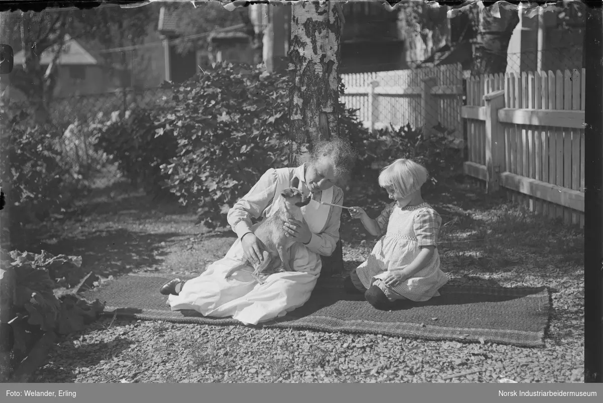 Kvinne og barn sittende på teppe i hage. Kvinnen har en hund på fanget som barnet gir noe til. Elevheimen i bakgrunnen.