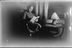 Kvinne sittende i stue og lese bok. Heklede gardiner og blen
