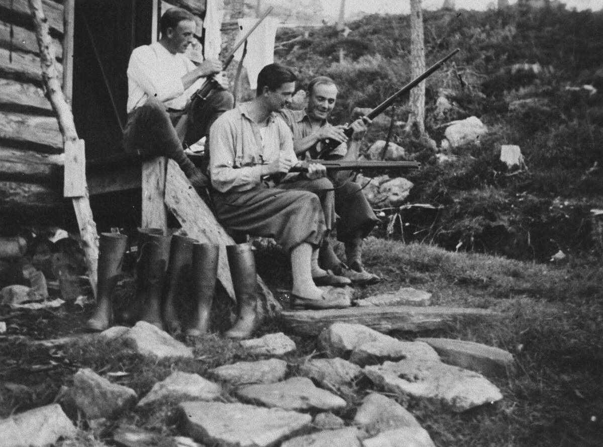 3 menn sitter ved inngangspartiet til ei hytte og pusser jaktgeværene sine.