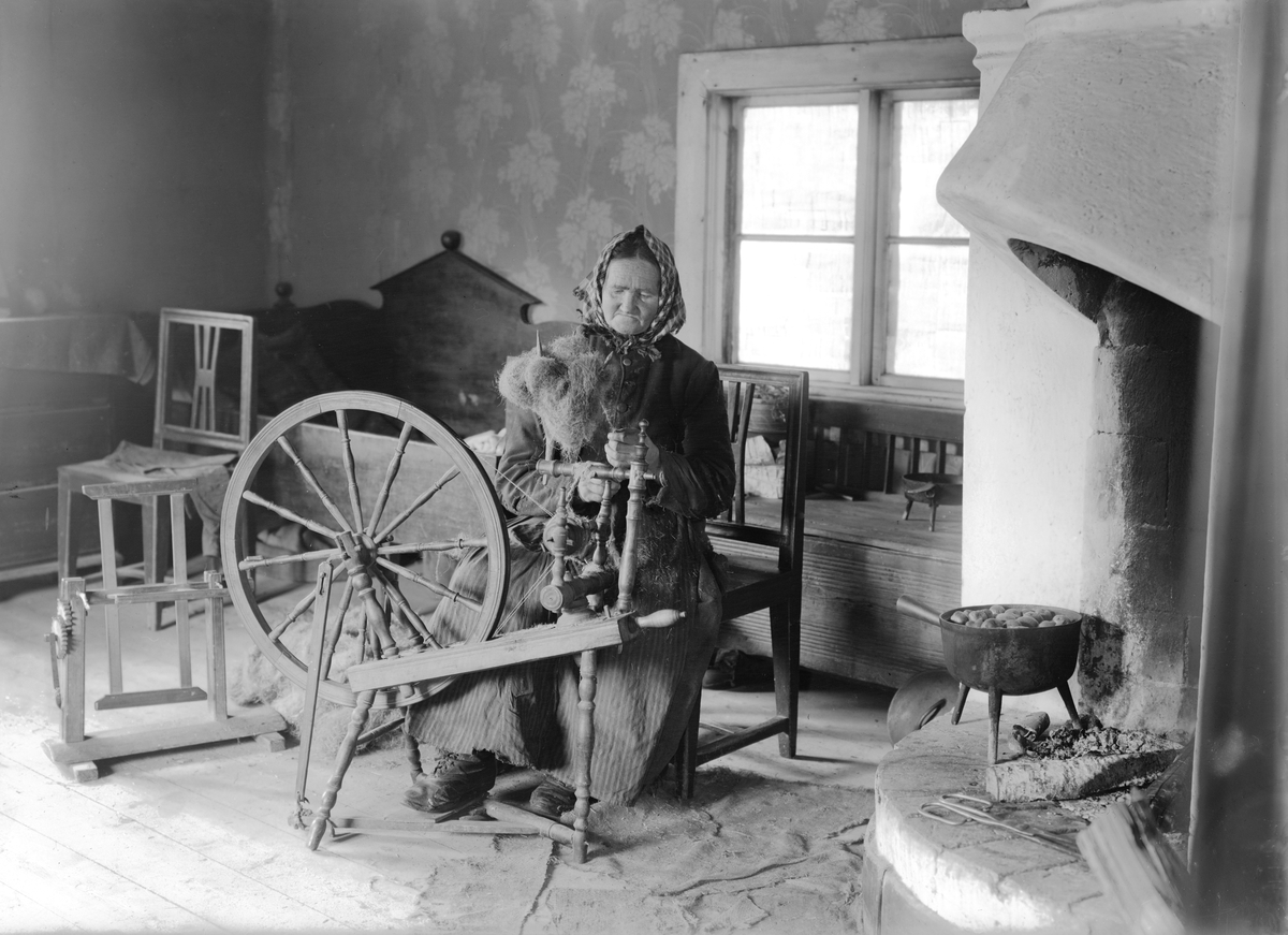 Interiör från backstugan Sjöhult i Svinhults sn. Vid spinnrocken sitter Karolina Eriksdotter. Vid tiden för bilden bor hon i stugan tillsammans med sin bror Johan Peter. Hon avled på ålderdomshemmet 1928.