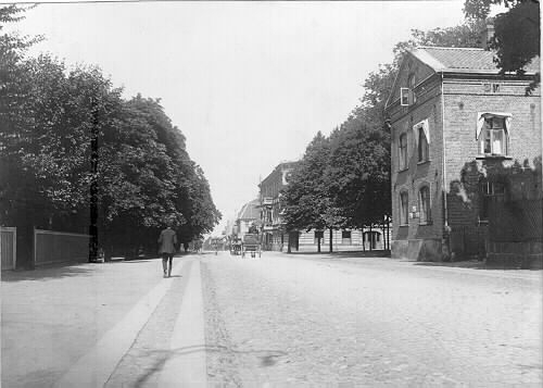 Vykort, Västra Vallgatan i Varberg mot norr med Jobsons magasin till höger med Brunnsparken på andra sidan. Till vänster i bild ligger kv Trädgården. Fototid: 1910-tal.