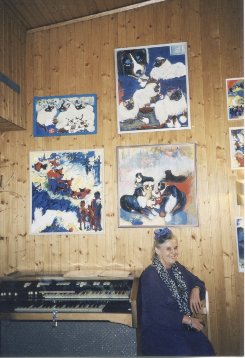 Hantverksutställning i Missionskyrkan, cirka 1990. Gertrud Dahlman sitter på en stol. Bakom henne hänger målade tavlor.