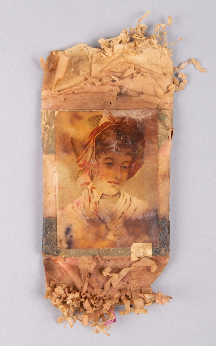 Julgranskaramell. Platt, rosafärgat silkespapper med fransar. Bild av ung flicka. Innehåller konfekt. Från 1930-talet enl. givaren.