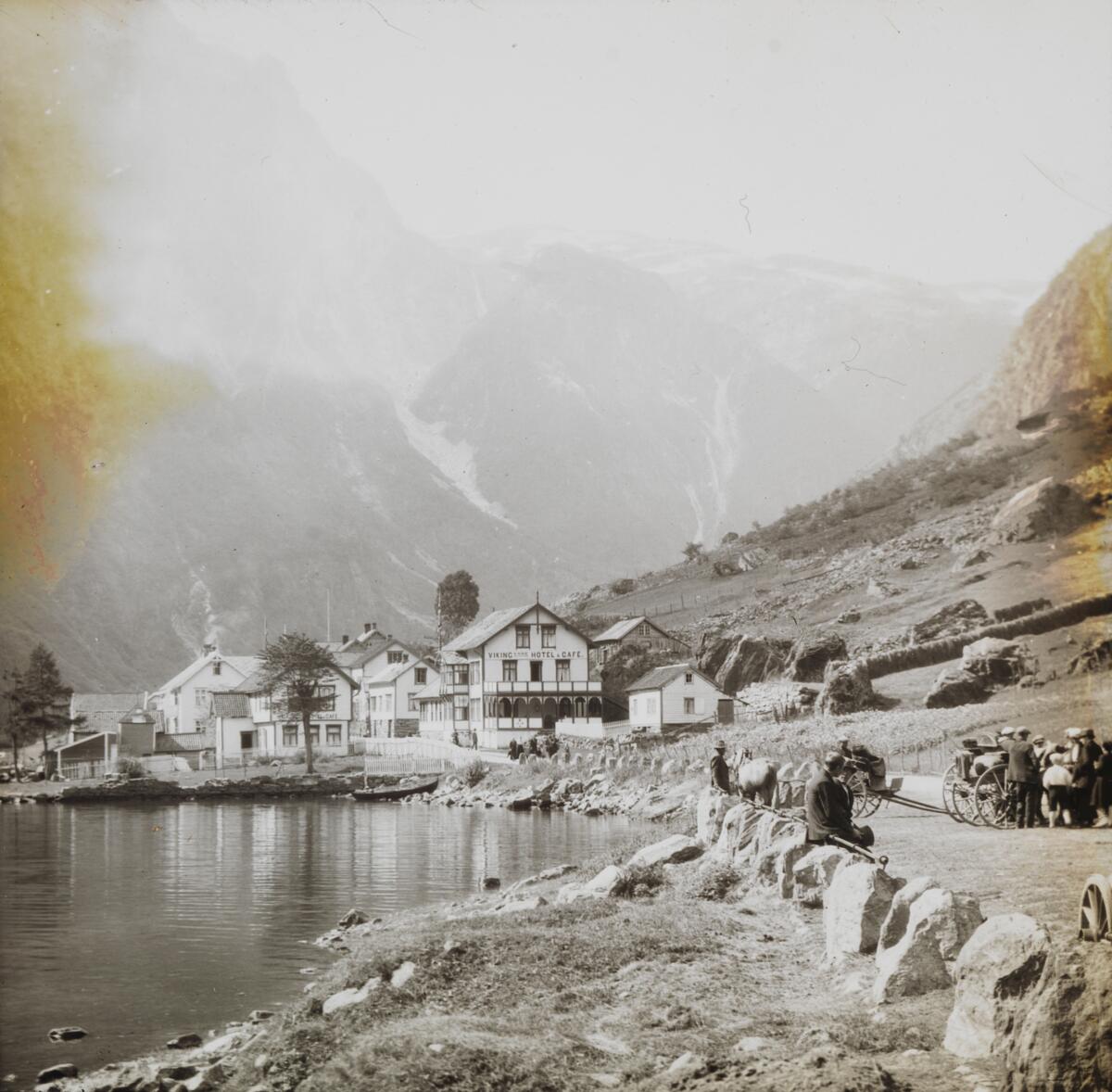Dias. En gruppe trehus i sveisterstil ligger på et nes i en fjord omgitt av bratte fjell. På veien i forgrunnen står en gruppe kvinner og menn, antakelig passasjerer fra passasjerskipet "Stavangerfjord". 