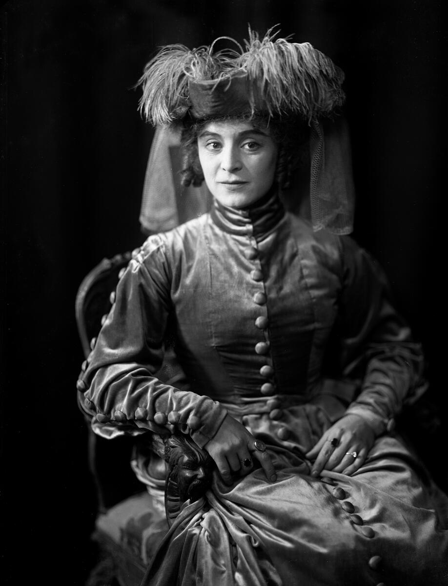 Portrett av skuespilleren og sceneinstruktøren Gerda Ring. Hun poserer sittende i en kjole og en stor hatt med fjær.