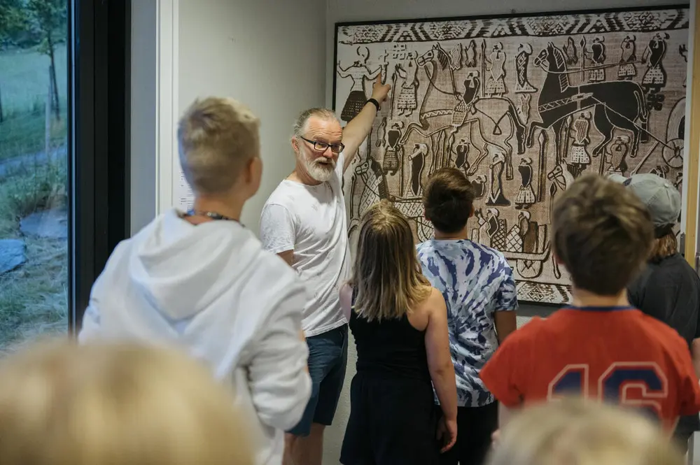 En mann peker opp på et bilde mens han forteller. Rundt han står flere barn i skolealder. De står med ryggen til kamera.