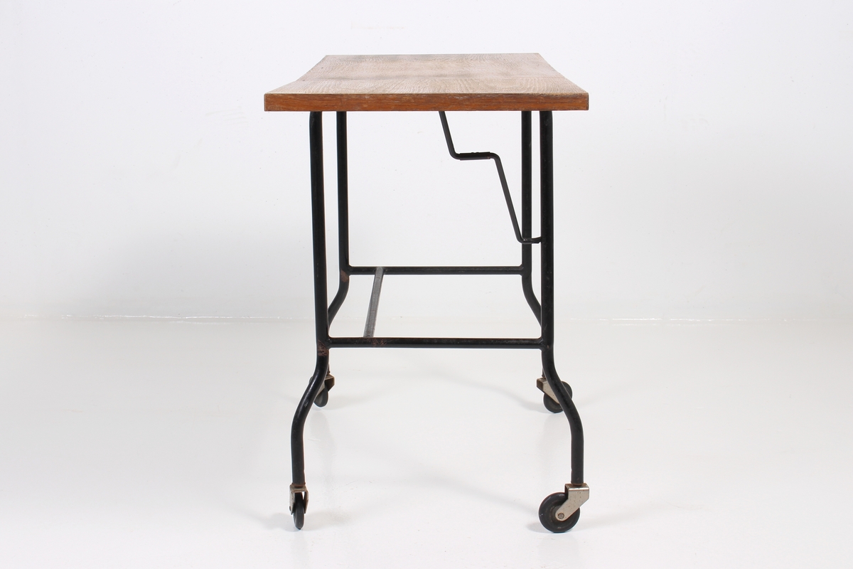 Lite avlastningsbord med finert bordplate, ramme av metallrør, og fire hjul. Bordplaten har en hengslet klaff som kan legges ned.