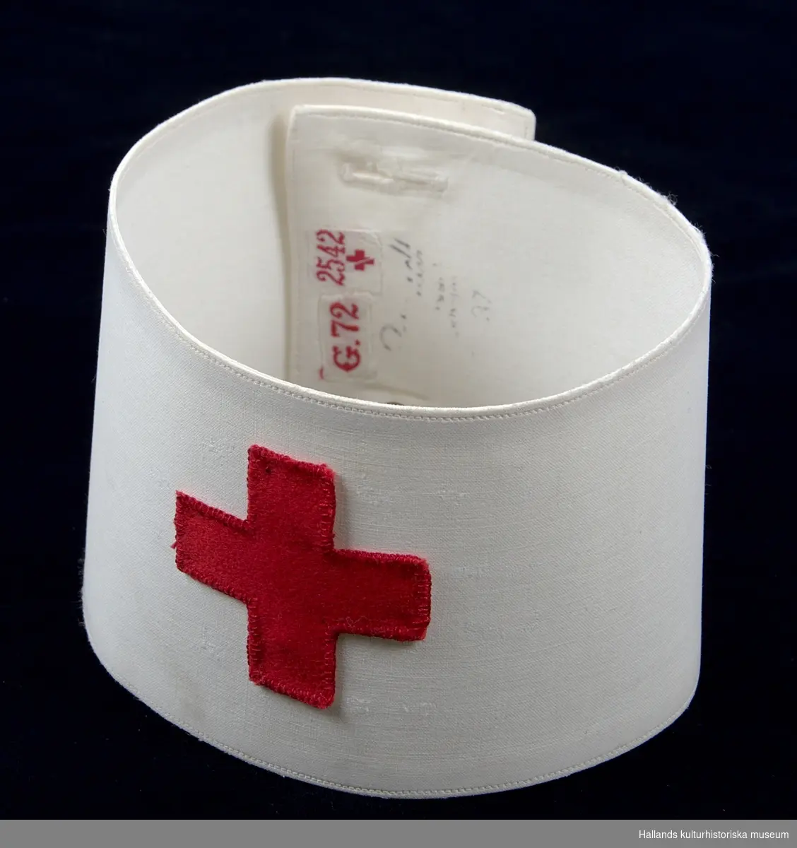 Armbindel till sjuksköterskeuniform. Tillverkad av manglad och pressad vit bomullsväv i tuskaft. Armbindeln bär Röda Korsets symbol i rött bomullstyg. Hålls ihop med två manschettknappar, varav den ena saknas. På insidan finns två etiketter (vit bakgrund med röd text): "G.72" och "2542" med ett rött kors. På insidan finns även en svårtydd stämpel i svart bläck (se foto)