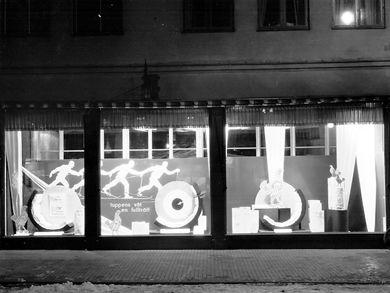 Bild tagen på kvällen i skyltfönstren hos Ekelöf & Svensson AB.
1872 etablerade fröknarna Hilda Ekelöf och Carolina Svensson en detaljaffär i manufaktur. Vacklande hälsa gjorde att firman 1897  överläts till Wilh. Welin som skapade nya avdelningar, till exempel flaggtillverkningen (1900) och en syatelje för klänningar (1905) samt en modeaffär (1914) där bland annat Josefin Baker gick modell.  Sommaren 1999 lades verksamheten ner på grund av olönsamhet.
Källa: Nyblom-Svanqvist, Näringsliv i Värmland, 1945 och Värmlands Folkblad.