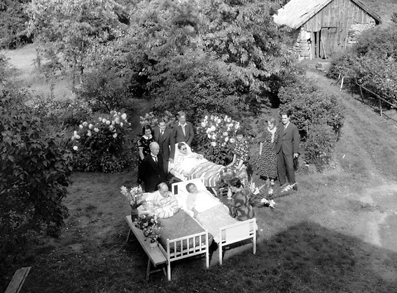 En grupp människor i trädgård varav tre ligger i sängar.