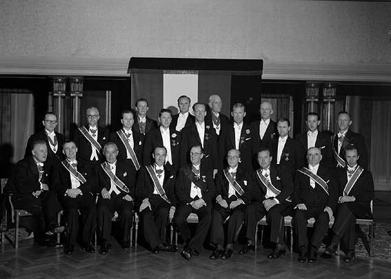 Större grupp män med ordensband.
Fotografens ant: Herr Lindqvist. Trikåfabriken.
Företaget grundades 1914 och då byggdes en fabrik upp på Herrhagen i Karlstad. I början tillverkades enbart bomullstrikå men efter några år togs även ylle och silke upp i produktionen samtidigt som fabriken utökades med eget färgeri och blekeri. 1937 inköpte bolaget Dala Väveri AB, Tidafors och Molkom som drevs som under namnet AB Värmlands Trikåfabrik avdelning B. Fabrikerna var mycket moderna med bland annat glastak för att tillvarata dagsljuset och högtalaranläggningar för att genom musik ge de anställda stimulans i arbetet.
Källa: Nyblom-Svanqvist, Näringsliv i Värmland, 1945.