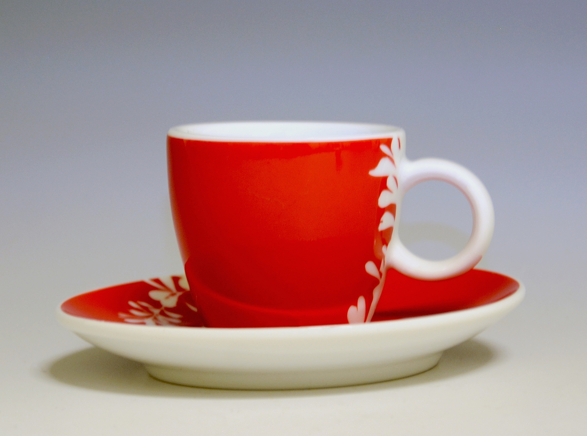 Espressokopp av porselen, med rund hank. Rød heldekkende dekor med utsparte blomster i hvitt.
Modell: Barista, forgitt av Poul Jensen.
Dekor: Red Kenya
