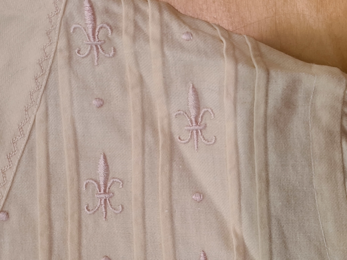 Pyjamas i tunn flanell. Långärmad och långbent. Lucka finns på baksidan. Försedd med två fickor. Dekoration med franska liljor. Vågigt broderat avslut på benen.