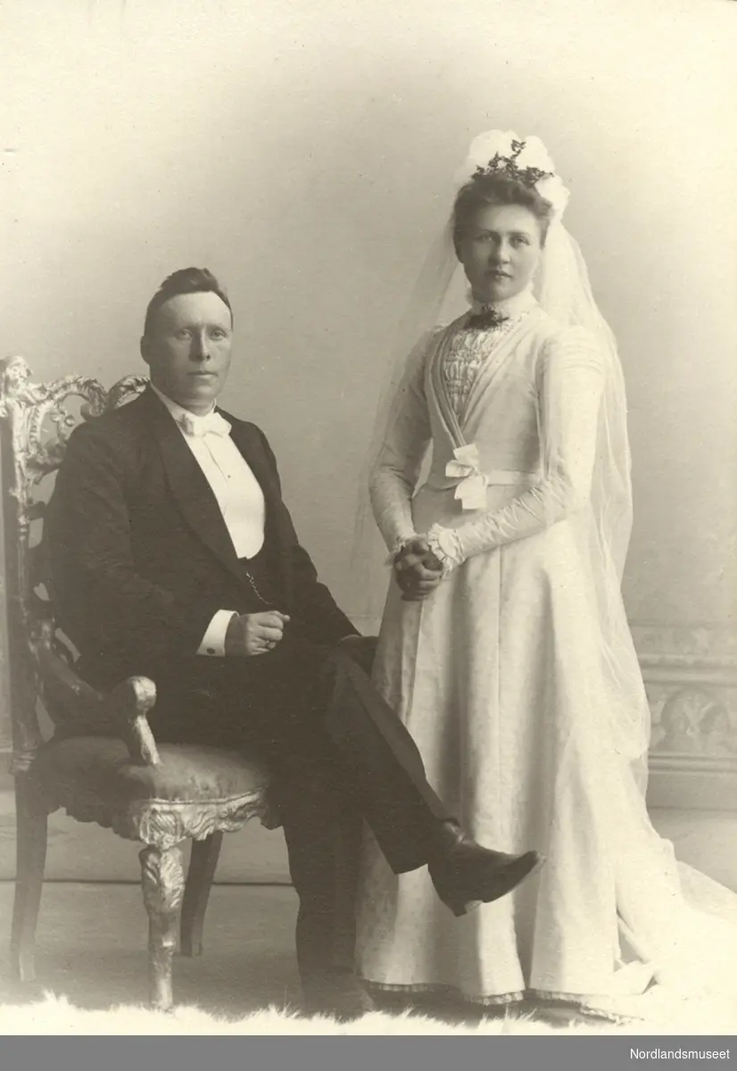 Sogneprest Nilsen Roald med sin Brud. Han sitter i en stol t.v. iført en pen dress, hun står t.h. i hvit brudekjole og slør, bildet tatt hos fotograf.