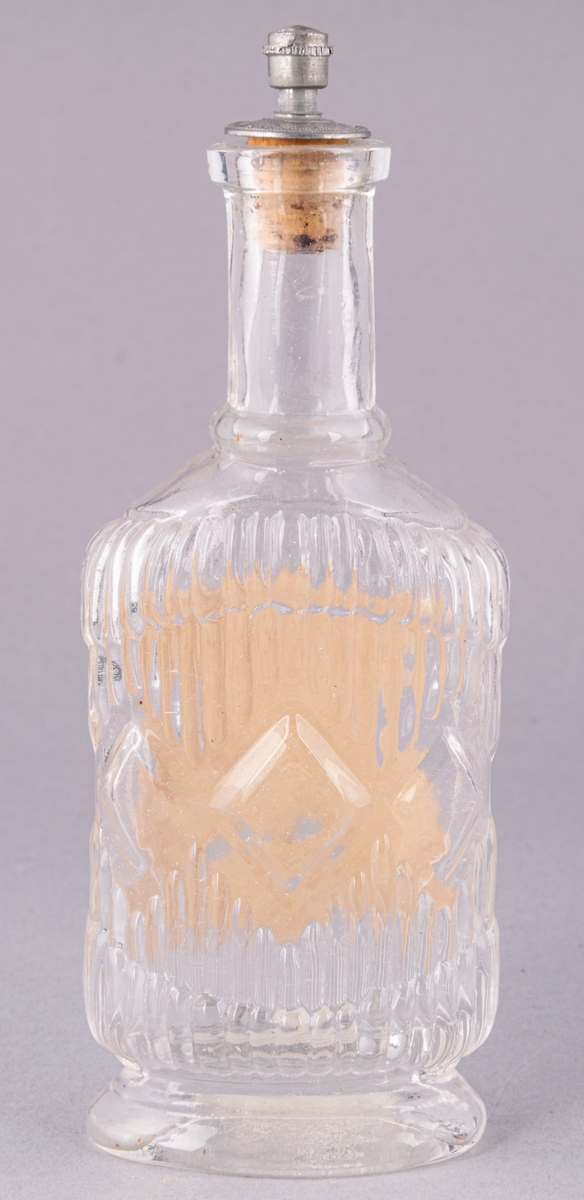 Parfymflaska, med glasknopp och gult band runt knoppen. Etikett med blomma och två ansikten.