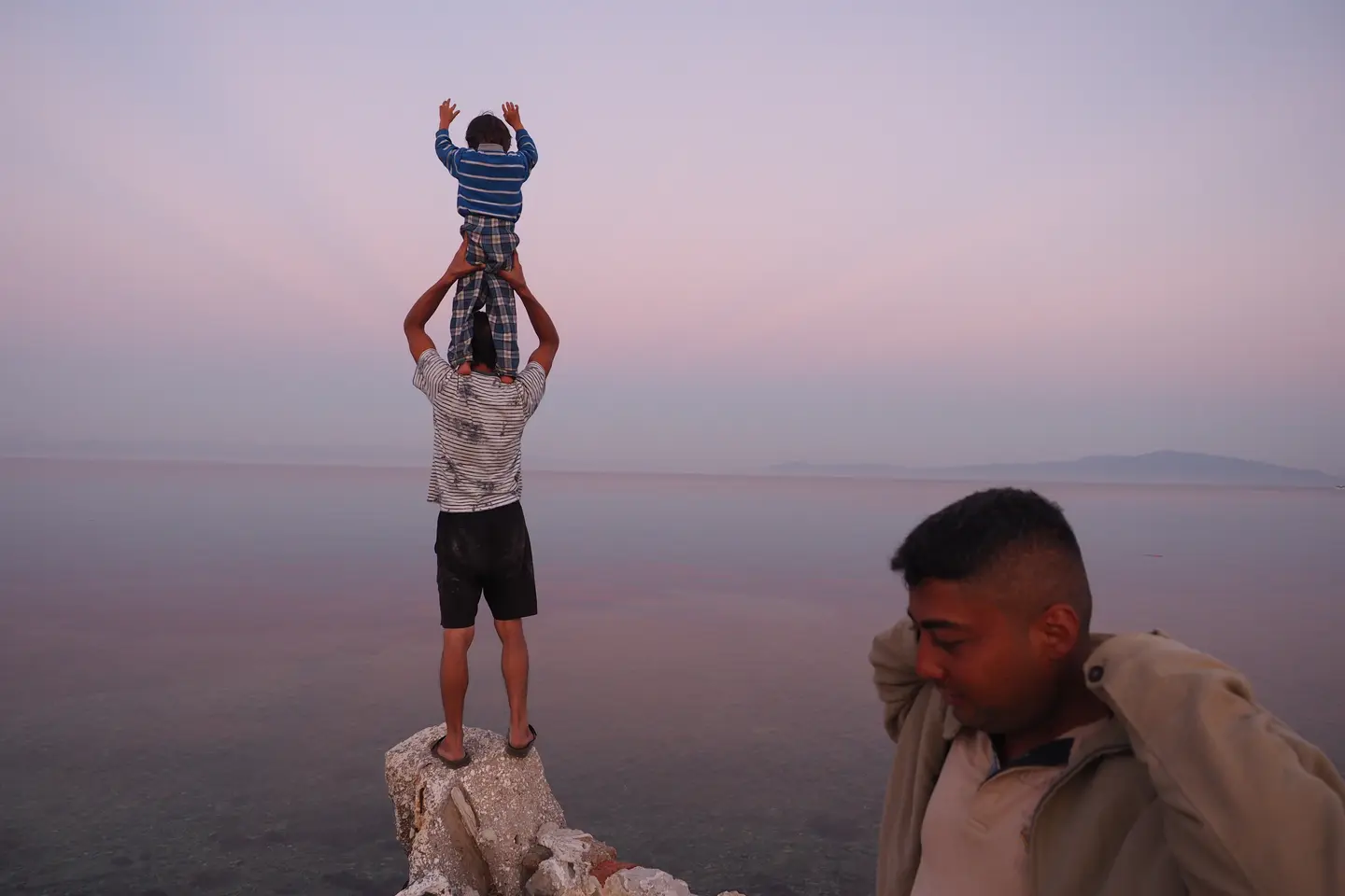 En stille kveld på stranden ved Pikpa med utsikt mot Tyrkia. En far står med sønnen på skuldrene og en annen person står i forgrunnen.